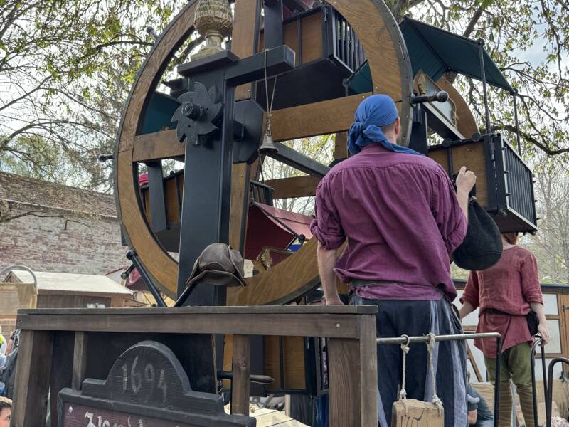 Ein mittelalterliches Holzriesenrad mit einer Handkurbel und mittelalterlich gekleideten Personen.