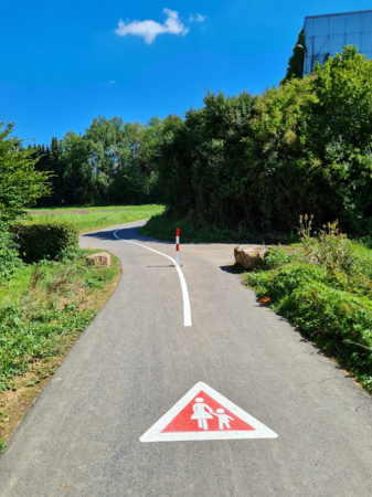 Ein Feldweg ist mit einem Hinweis auf Fußgänger markiert. Im Hintergrund wird der Weg durch eine Bodenmarkierung getrennt.