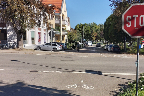 An einer Kreuzung steht ein Stop-Schild. Die Straße ist mit einem Fahrrad-Piktorgramm markiert. Im Hintergrund sind parkende Autos und Menschen auf Fahrrädern.