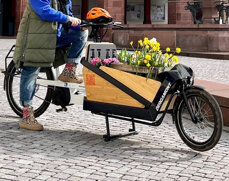 Eine Frau transportiert Blumen mit einem Lastenrad. Im Hintergrund befindet sich das Rathaus und ein Brunnen.