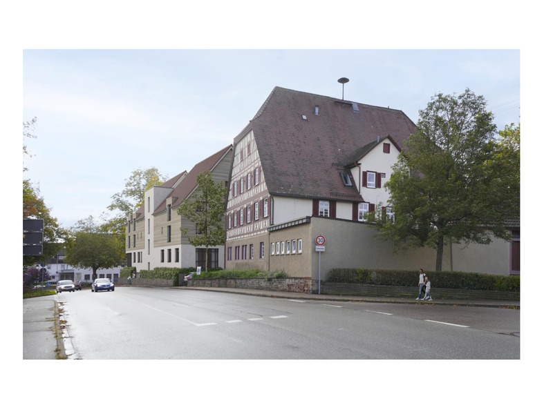 Eine Fachwerkhausfassade mit großem Dach, flankiert von Bäumen und einer Straße mit Gehsteig, an der zwei Personen entlanggehen.
