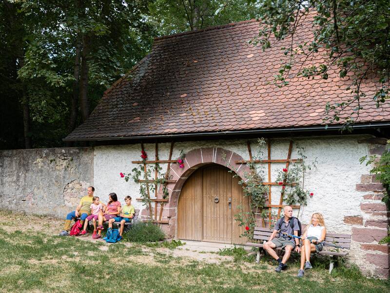 Eine Familie und ein Paar ruhen sich auf Bänken vor einem alten Haus aus.
