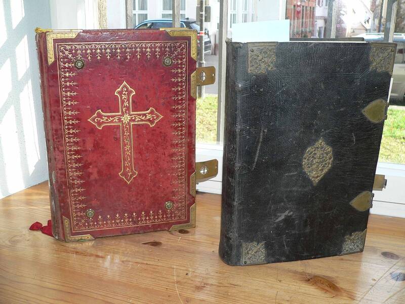 Zwei aufgestellte Bücher. Das linke Buch hat einen roten Einband mit einem Kreuz. Das rechte Buch ist schwarz.