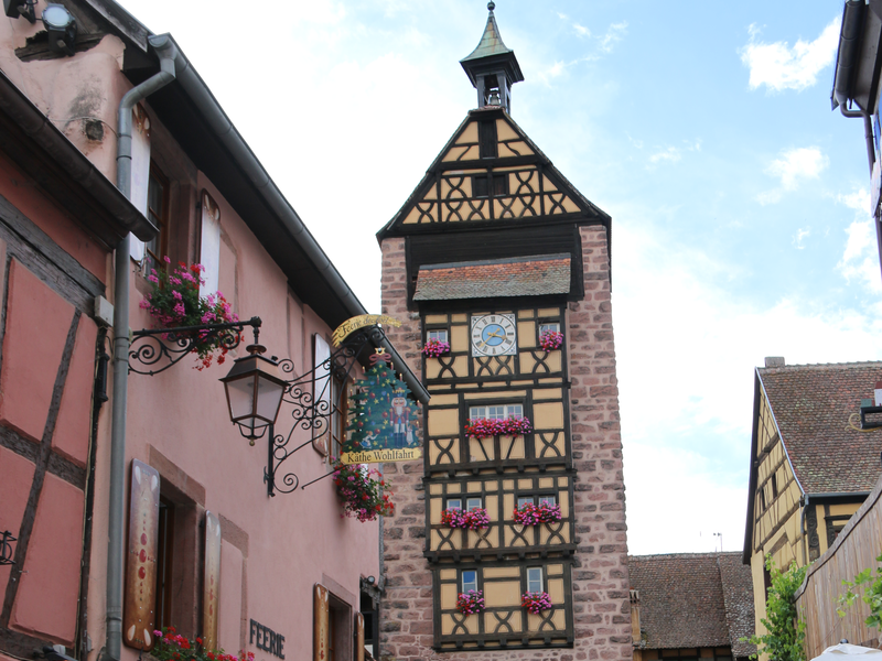 Menschen laufen durch die Altstadt von Riquewihr. Im Hintergrund ist der Dolder, das Wahrzeichen.