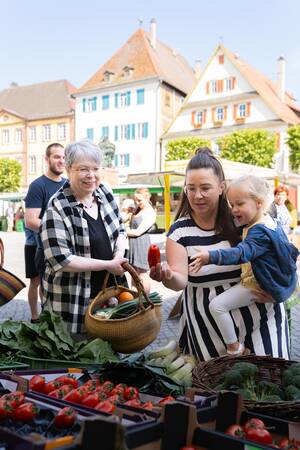 Mutter zeigt Kind Paprika am Wochenmarktstand mit lächender Seniorin nebenan