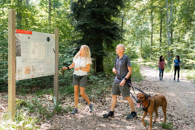 Ein Ehepaar mit Hund steht an einem Infoschild, im Hintergrund laufen zwei Frauen