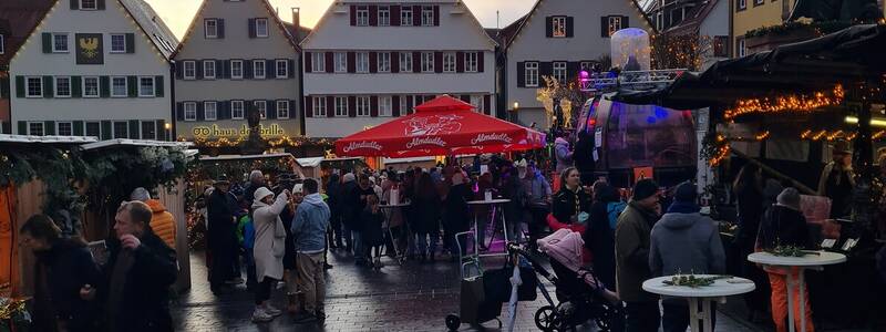 Eine belebte Marktszenerie in einer Altstadt mit Menschen, die zwischen festlich dekorierten Ständen flanieren, während der Himmel im Hintergrund farbig leuchtet.