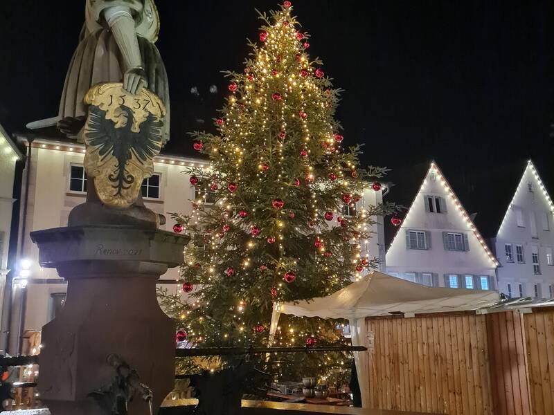 Ein großer, festlich geschmückter Weihnachtsbaum steht auf einem Platz neben einer Statue und traditionellen Gebäuden, die im Dunkeln von Lichtern beleuchtet werden.