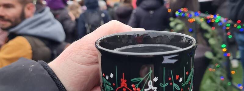 Eine Hand hält einen weihnachtlich dekorierten Becher auf einem belebten Weihnachtsmarkt, während Menschen mit Regenschirmen im Hintergrund zu sehen sind.