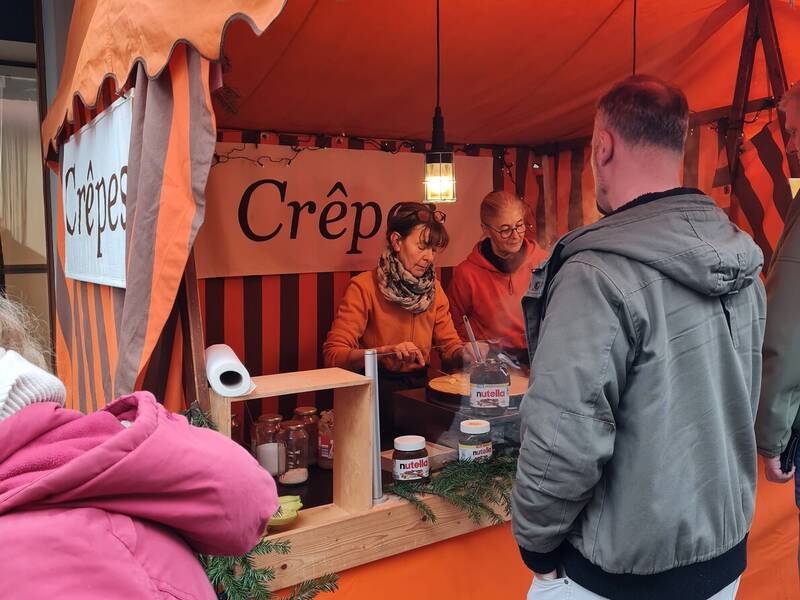 Ein orangefarbener Crêpes-Stand auf einem Markt mit zwei Verkäuferinnen, die Crêpes zubereiten, während Kunden davor warten.
