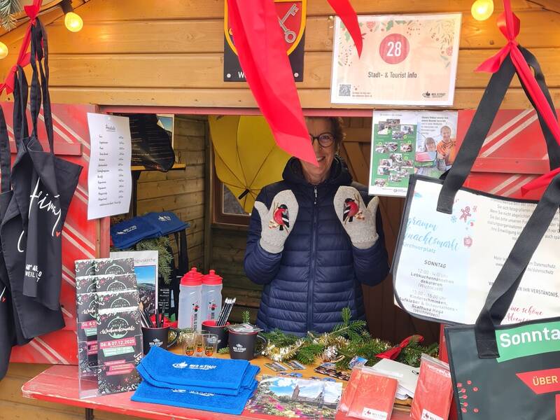 Eine Frau steht in einem festlich dekorierten Holzstand mit roten Schleifen und verschiedenen Informationsmaterialien sowie Souvenirs auf einem Tisch.