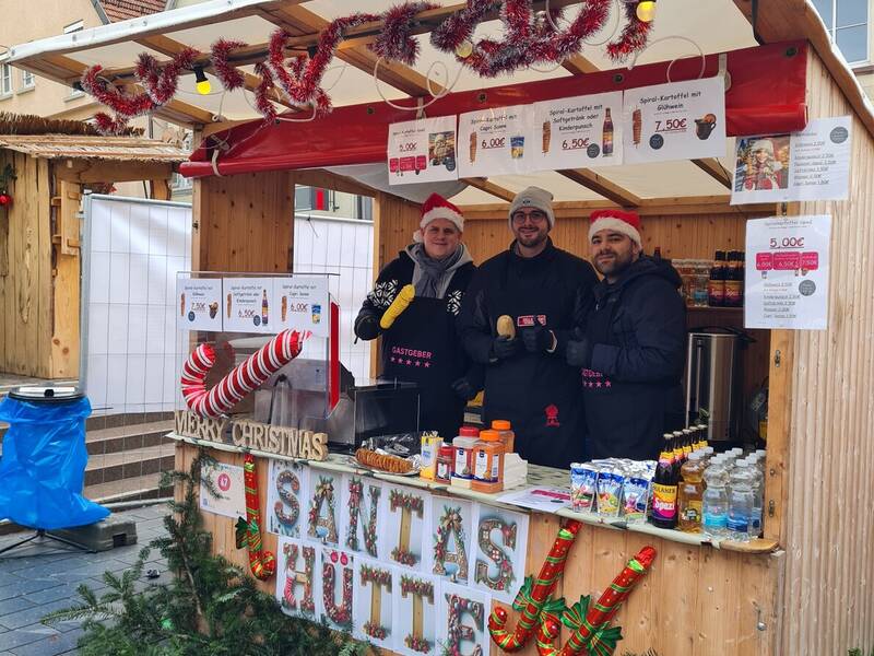 Drei Personen stehen in einer festlich geschmückten Holzhütte mit dem Schild "Santas Hütte" auf einem Weihnachtsmarkt und bieten Speisen und Getränke an.