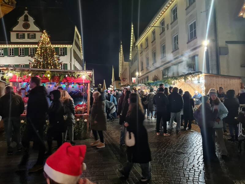 Eine belebte Szene eines Weihnachtsmarkts bei Nacht mit einem großen beleuchteten Weihnachtsbaum im Hintergrund und geschäftigen Ständen, umgeben von Menschenmengen.