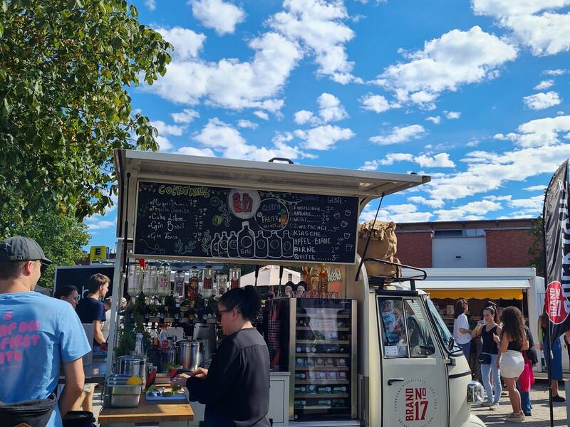 Eine mobile Bar auf einem kleinen dreirädrigen Fahrzeug bietet unter blauem Himmel mit weißen Wolken verschiedene Getränke an, während Menschen davor stehen und bestellen.