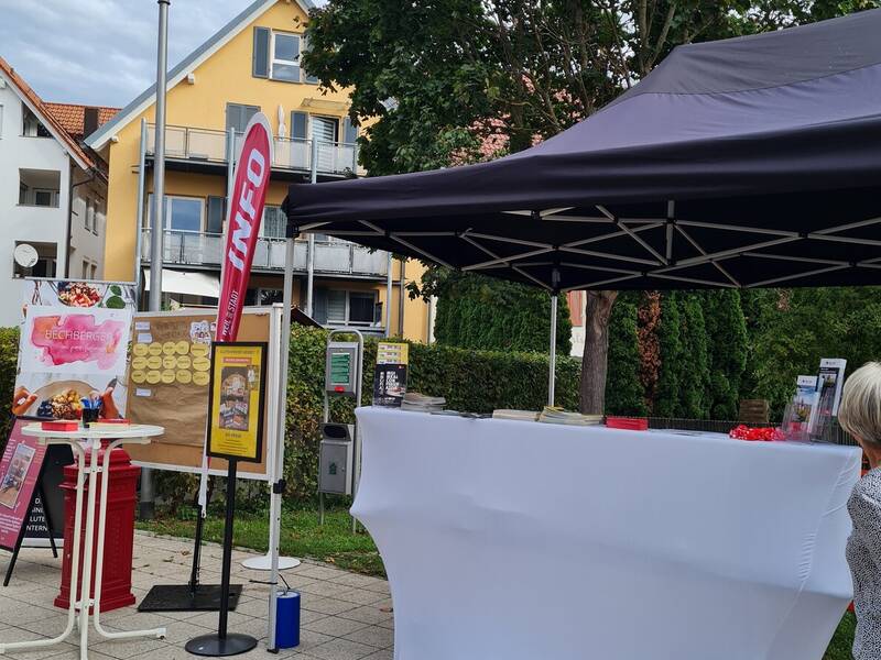 Ein Pavillon mit einem weißen Tisch und Infomaterialien steht auf einem gepflasterten Platz vor einem gelben Haus und einem Baum, daneben ist ein Banner mit der Aufschrift "INFO" und eine Tafel mit Notizen zu sehen.