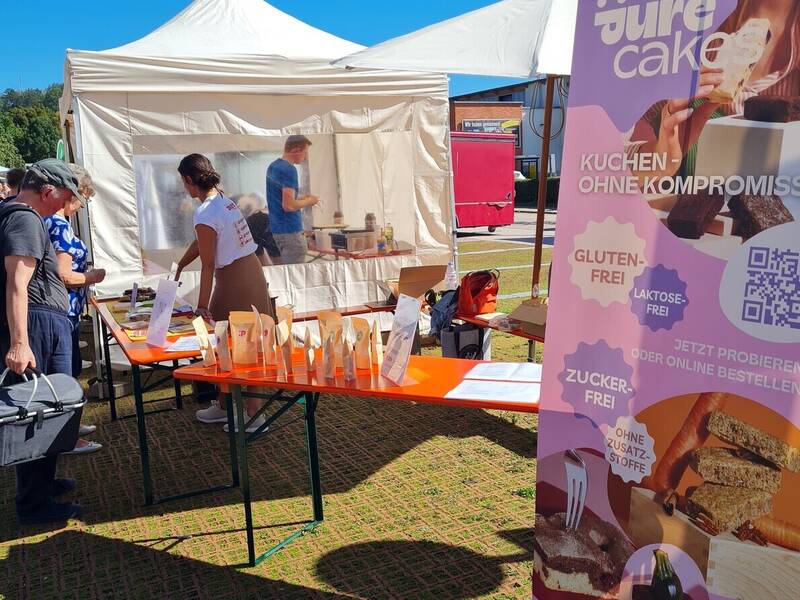 Ein Marktstand mit orangefarbenen Tischen bietet glutenfreie, laktosefreie und zuckerfreie Produkte unter einem weißen Zelt an, während ein Werbebanner Informationen darüber präsentiert.