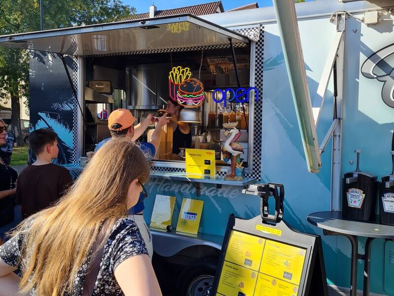 Menschen stehen an einem Foodtruck mit einem Menü im Vordergrund an, der Pommes und Hamburger anbietet.
