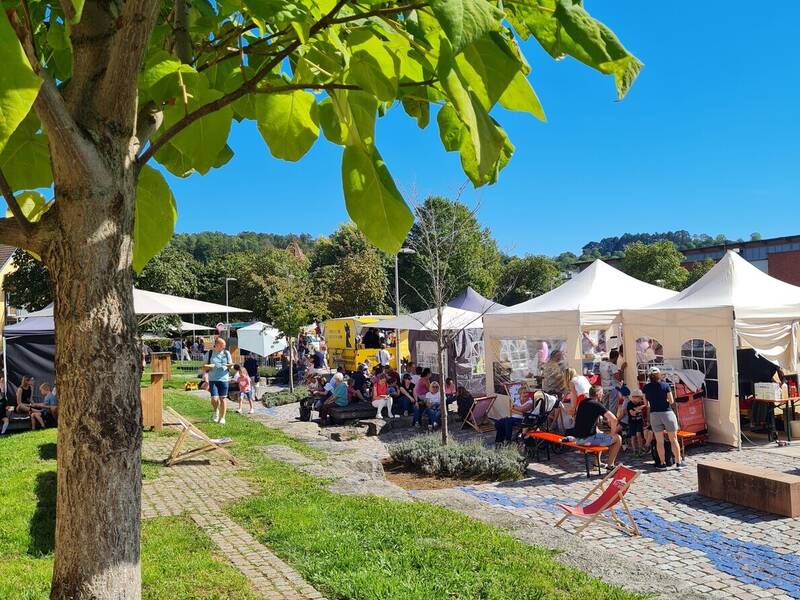 Auf dem Bild ist ein lebhafter Markt bei sonnigem Wetter zu sehen, mit Zelten, Ständen und vielen Menschen, die sich auf einem pflastersteingedeckten Platz entspannen.