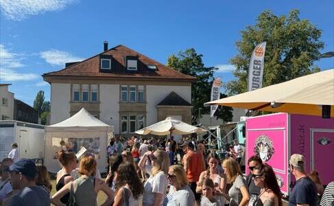 Eine große Gruppe von Menschen steht bei sonnigem Wetter in einer Schlange vor Essensständen auf einem sommerlichen Straßenfest.