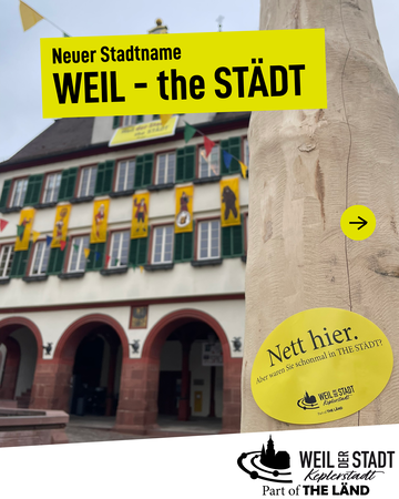 Ein Schild mit der Aufschrift "Neuer Stadtname WEIL - the STÄDT" hängt vor dem Rathaus mit bunten Fahnen.