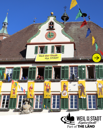 Ein gelber Banner mit der Aufschrift "Weil der Stadt the STÄDT" hängt über einer Reihe farbenfroher Figurenbanner an Fenstern des Rathauses, vor einem klaren blauen Himmel.