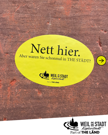 Ein gelbes ovales Schild auf einer rötlichen Steinoberfläche trägt die Aufschrift "Nett hier. Aber waren Sie schonmal in THE STÄDT?" mit einem Logo und Pfeil.