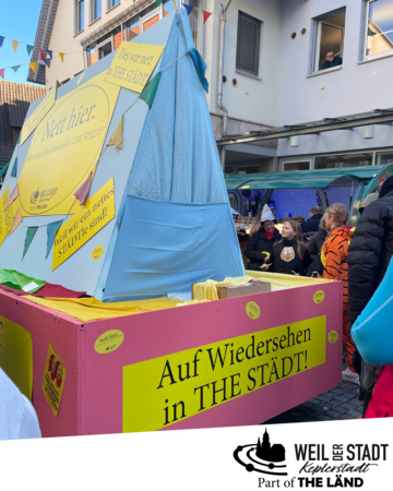 Ein bunter Fasnetswagen mit dem Schriftzug "Auf Wiedersehen in THE STÄDT!" ist von verkleideten Menschen umgeben.