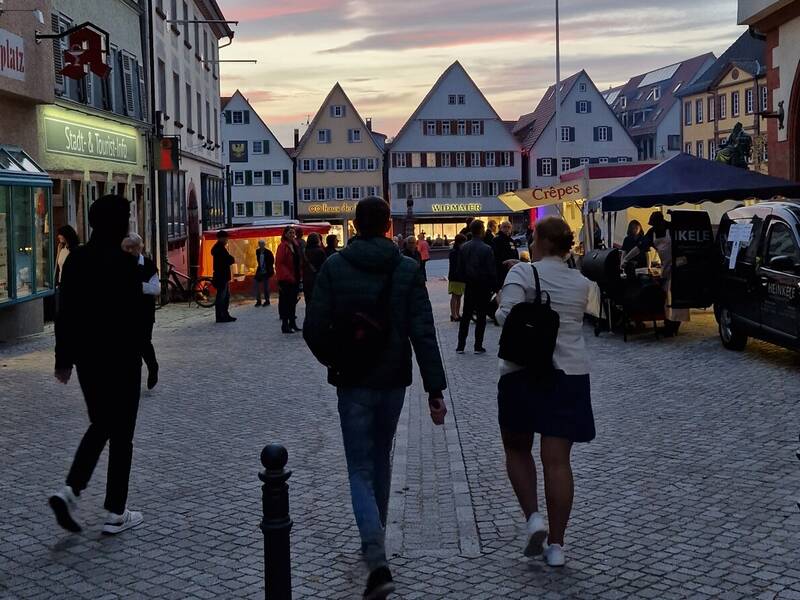 Ein Kopfsteinpflasterweg führt durch eine belebte Straße mit Geschäften und Ständen, während Menschen in Richtung der im Hintergrund sichtbaren, spitzgiebeligen Häuser spazieren, unter einem Himmel mit malerischem Sonnenuntergang.