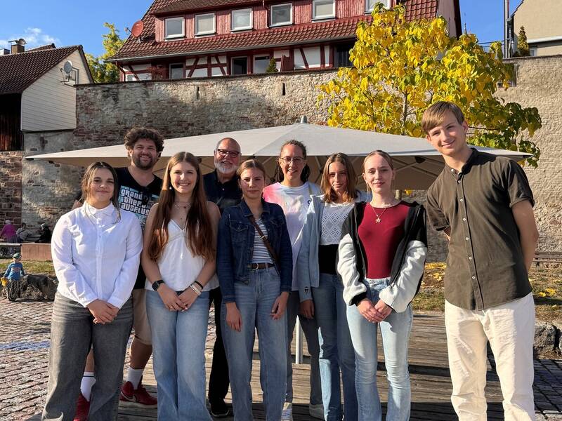Eine Gruppe von neun Personen steht auf einem Holzdeck mit der Stadtmauer und einem gelben Baum im Hintergrund. Es handelt sich um die Mitglieder des Jugendbeirats und Vertreter der Verwaltung.