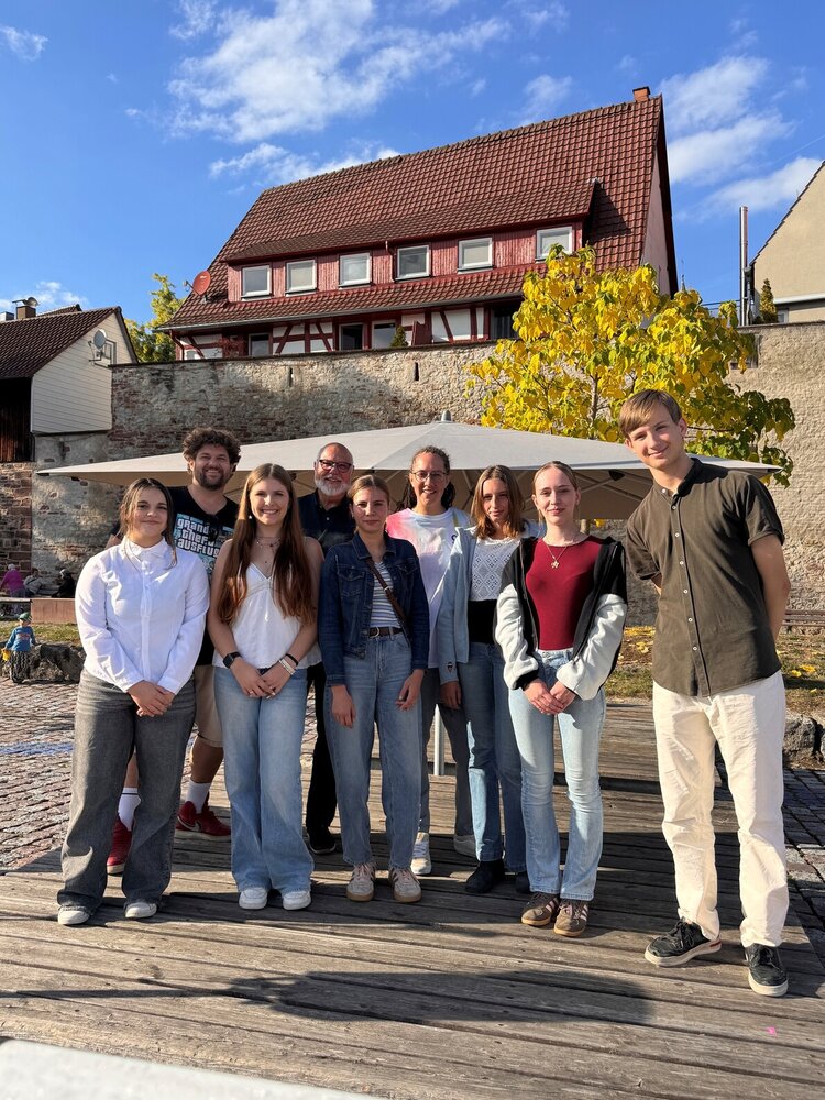 Eine Gruppe von neun Personen steht auf einem Holzdeck mit der Stadtmauer und einem gelben Baum im Hintergrund. Es handelt sich um die Mitglieder des Jugendbeirats und Vertreter der Verwaltung.