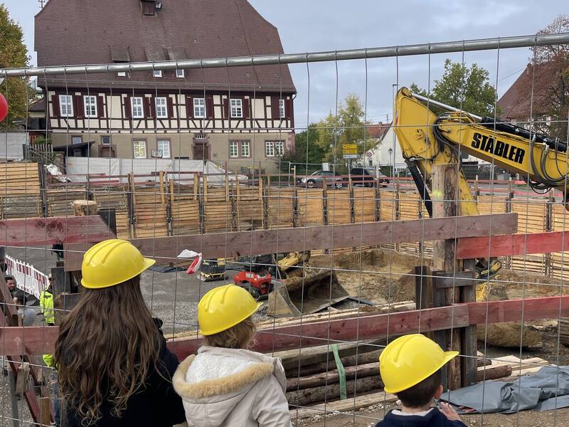 Drei Kinder mit gelben Schutzhelmen stehen vor einem Bauzaun und sehen auf eine Baustelle mit einem Bagger, während im Hintergrund ein großes Fachwerkhaus zu sehen ist.
