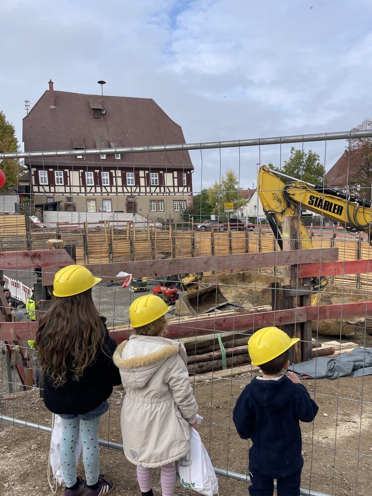 Drei Kinder mit gelben Schutzhelmen stehen vor einem Bauzaun und sehen auf eine Baustelle mit einem Bagger, während im Hintergrund ein großes Fachwerkhaus zu sehen ist.