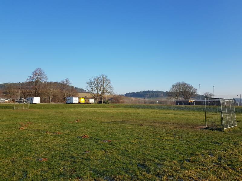 Eine grüne Rasenfläche mit einem leeren Tor im Vordergrund, umgeben von einigen Bäumen und Wohnwagen im Hintergrund unter blauem Himmel.