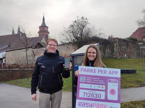 Zwei Verwaltungsmitarbeiter machen mit einem Smartphone und einem Schild in der Hand WErbung für die Park-App.