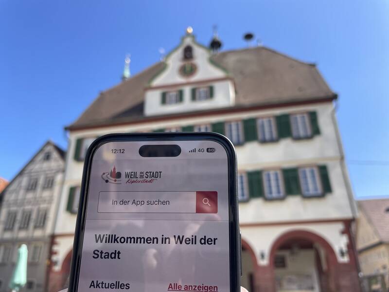 Ein Smartphone zeigt die geöffnete Bürger-App mit der Begrüßung "Willkommen in Weil der Stadt" vor dem Rathaus.