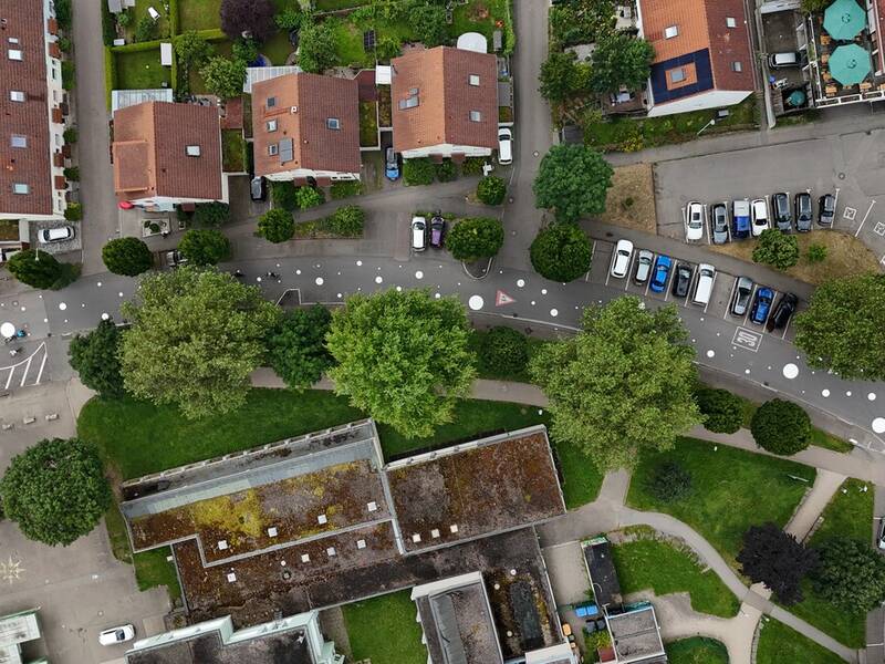 Eine Luftaufnahme zeigt eine Wohnstraße mit geparkten Autos, roten Ziegeldächern, Bäumen und einem umliegenden Wohngebiet.