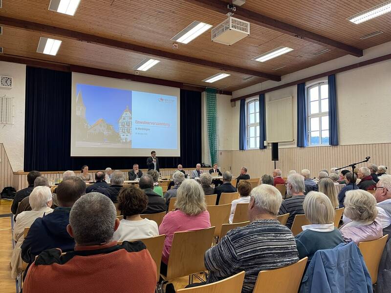 In einer Halle sitzen zahlreiche Menschen  blicken auf eine Leinwand mit der Aufschrift "Einwohnerversammlung in Merklingen".