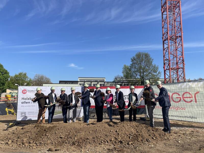 Eine Gruppe von Menschen mit Bauhelmen hält Spaten und steht vor einem Schild bei einer Spatenstichzeremonie auf der Baustelle der neuen Grundschule.