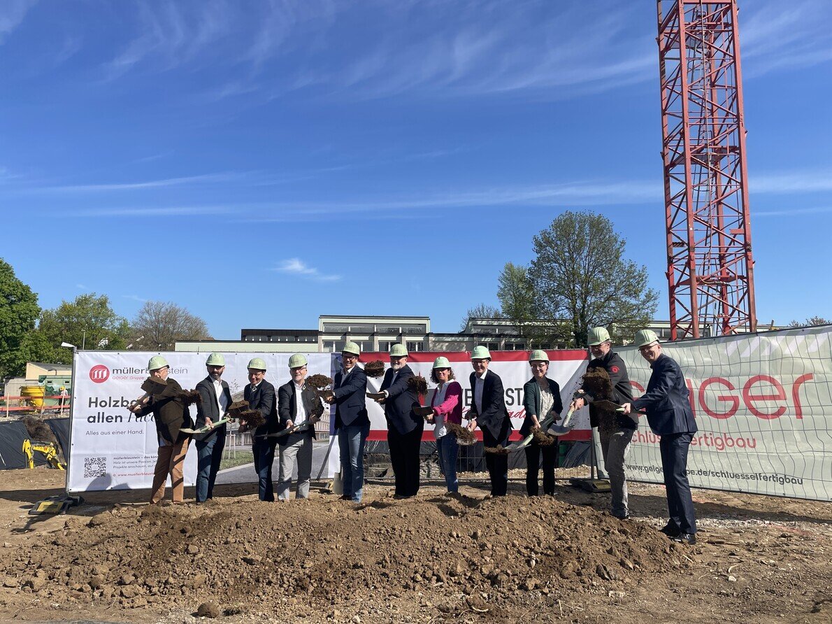 Eine Gruppe von Menschen mit Bauhelmen hält Spaten und steht vor einem Schild bei einer Spatenstichzeremonie auf der Baustelle der neuen Grundschule.