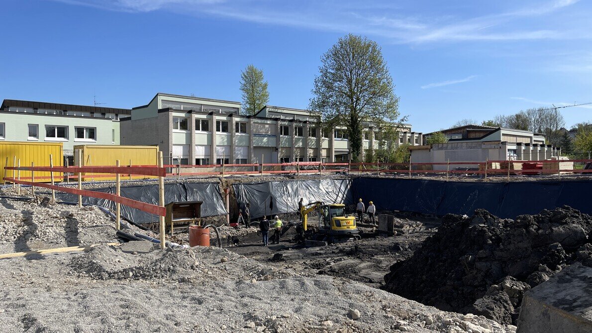 Auf einer Baustelle vor einem Schulgebäude arbeiten Bauarbeiter mit einem kleinen Bagger in einer großen Baugrube. 