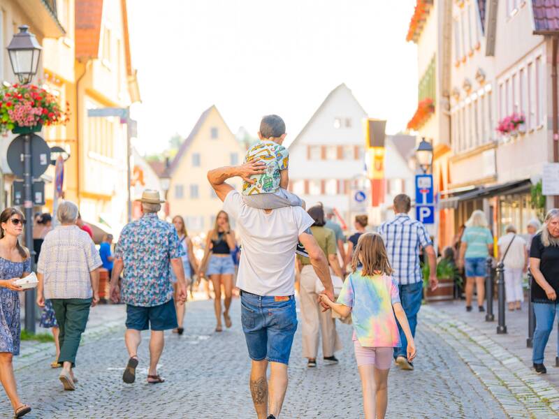 Eine belebte, sonnige Fußgängerstraße in der Altstadt von Weil der Stadt mit Menschen, die spazieren und Geschäfte auf beiden Seiten.