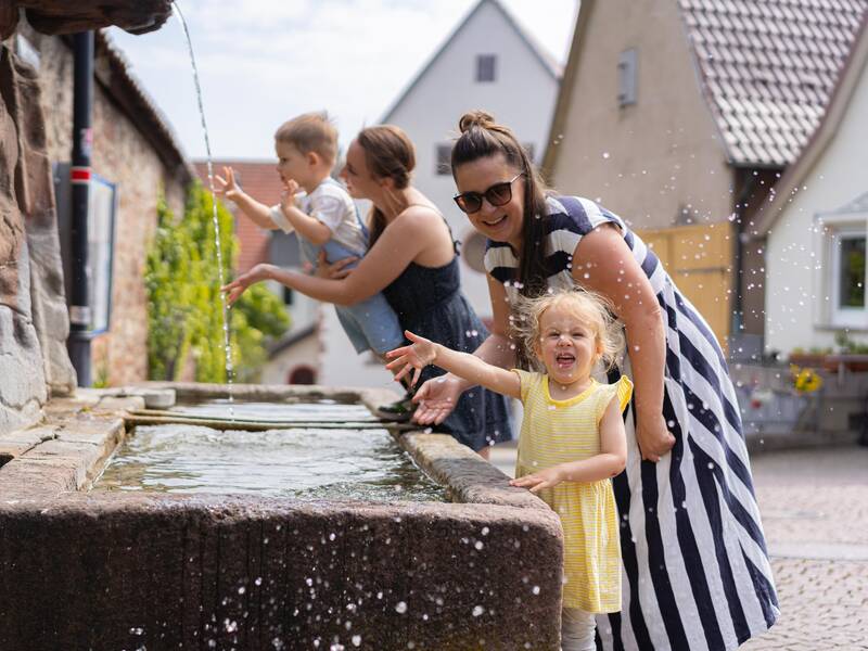 Zwei Frauen spielen mit ihren Kindern an einem Brunnen.