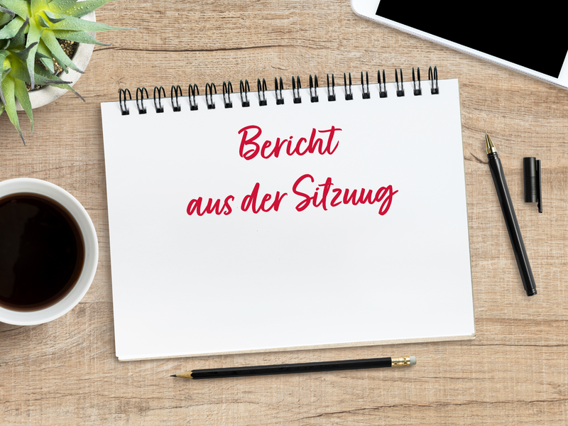 Auf einem Arbeitsplatz liegen ein Block, ein Handy und verschiedene Stifte. Auf dem Block steht "Bericht aus der Sitzung".