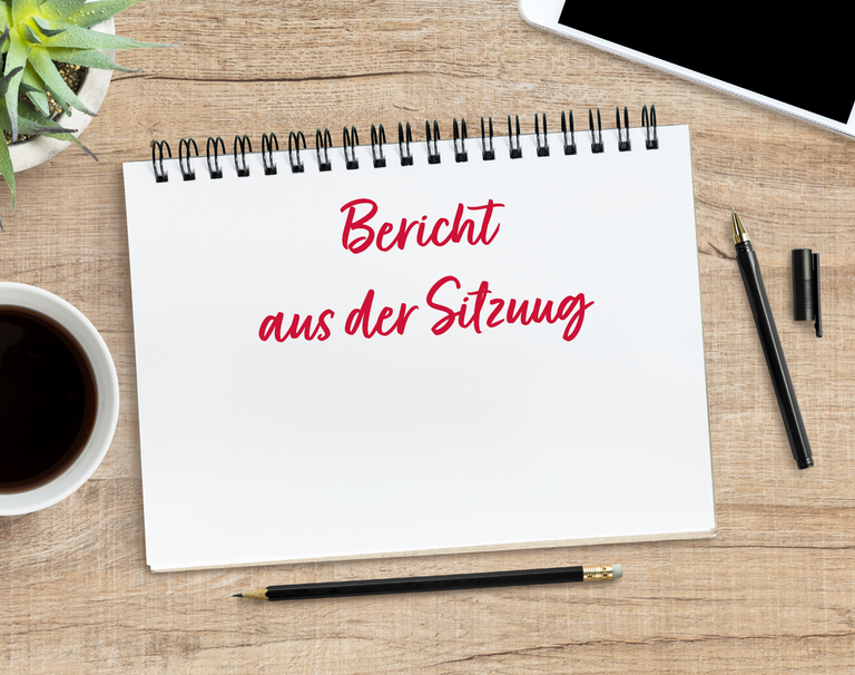 Auf einem Arbeitsplatz liegen ein Block, ein Handy und verschiedene Stifte. Auf dem Block steht "Bericht aus der Sitzung".