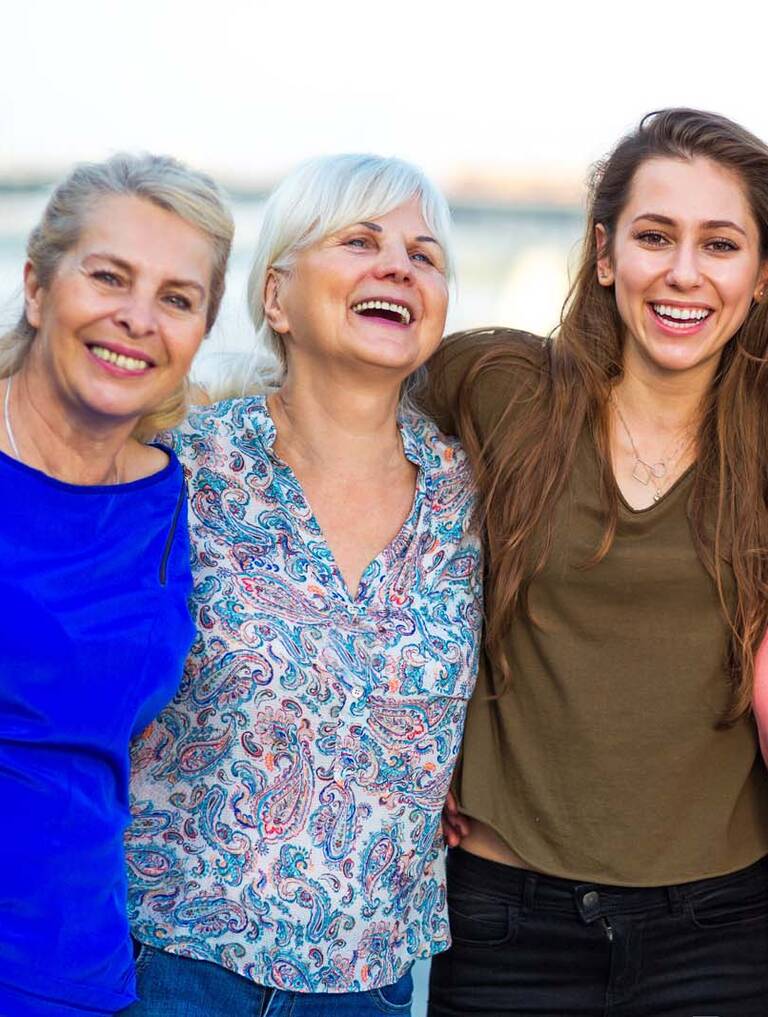 Eine Gruppe von vier Frauen unterschiedlichen Alters umarmen einander und lachen. Im Hintergrund ist ein Gewässer zu sehen.