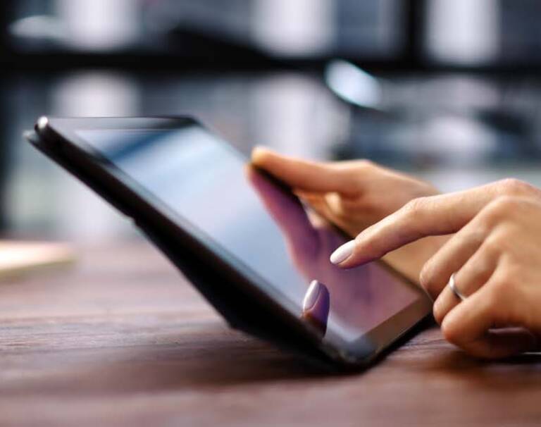 Zwei Hände halten ein Ipad auf einem Tisch. Links sind Blöcke und Stifte.
