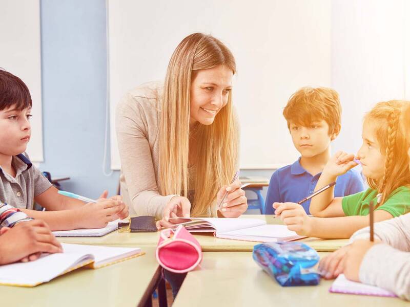 Eine lächende Schülerbetreuerin erklärt Kindern etwas an einem Tisch.
