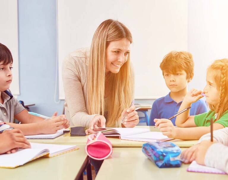 Eine lächende Schülerbetreuerin erklärt Kindern etwas an einem Tisch.