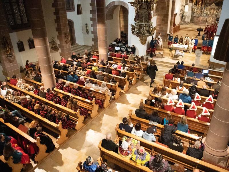 Eine bunte Gruppe von Menschen in fantasievollen Fasnetskostümen sitzen in einer Kirche und verfolgen eine lebhafte Narrensitzung.