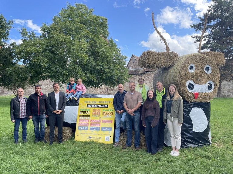 Eine Gruppe von Menschen steht vor einem großen, als Kuh gestalteten Heuballen und einem Schild, das für das „Weiler Herbst“ Festival wirbt.
